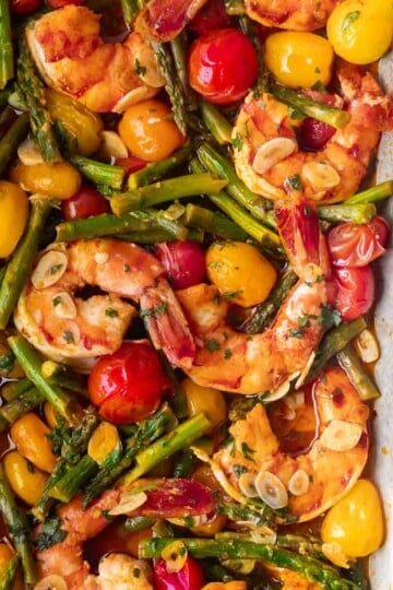 Sheet pan garlic shrimp with cherry tomatoes, asparagus, and garlic slices on a tray garnished with fresh herbs.