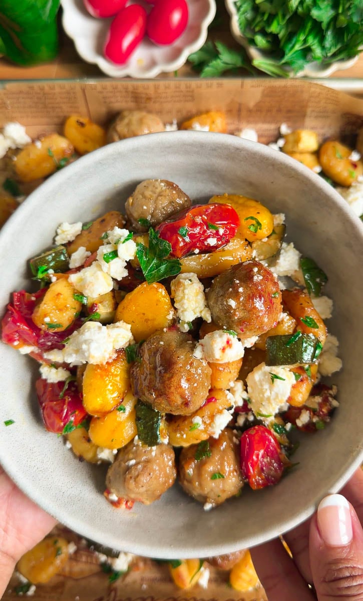 Sheet pan baked gnocchi with meatballs, cherry tomatoes, zucchini, feta, and herbs on a wooden table.