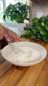 Person whisking flour in a white bowl on a wooden countertop.