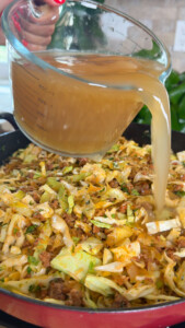 Pouring broth into a skillet with chopped cabbage and ground meat cooking.