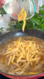 Pouring penne pasta into a steaming pot of broth on a kitchen stove.