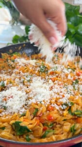 Hand sprinkling grated cheese over cooked orzo in a skillet.