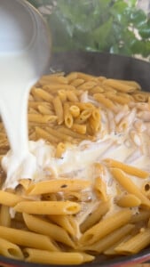 Pouring cream over penne pasta in a pan.