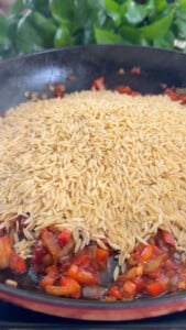 Orzo pasta with diced red peppers and onions cooking in a black skillet.