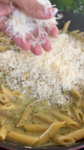Hand sprinkling grated cheese over pesto penne pasta in a pan.