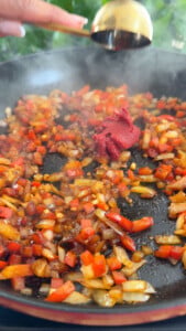 Sautéed onions and red tomatoes in a pan with tomato paste being added from a ladle.