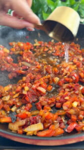 Hand pouring white wine into a skillet with sautéed diced red peppers and onions.
