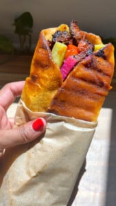 Hand holding wrapped shawarma with grilled meat, pickles, and vegetables.