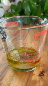 Glass measuring cup with layered oil, herbs, and liquid on a wooden table.