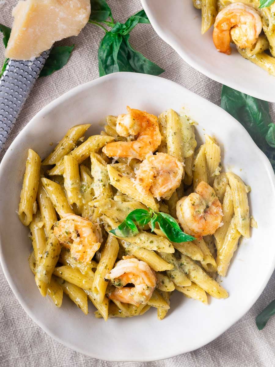 Pesto shrimp pasta in a white bowl, garnished with basil leaves, on a linen tablecloth with Parmesan cheese nearby.