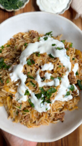 Rice dish with meat and herbs, topped with creamy white sauce, served in a white bowl on a wooden table.