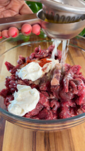 Squeezing lemon juice onto raw beef strips with yogurt and spices in a glass bowl.