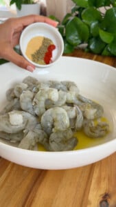 Bowl of raw shrimp with olive oil, hand holding small bowl with spices