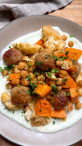 Bowl of roasted vegetables and meatballs with chickpeas on a creamy sauce, garnished with fresh herbs.