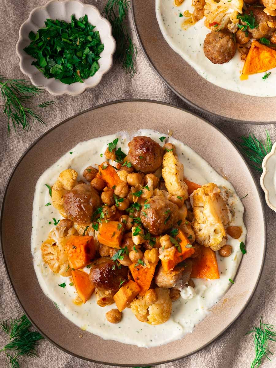 Sheet pan meatballs and veggies on creamy yogurt sauce, garnished with parsley, served on a beige plate with herbs.