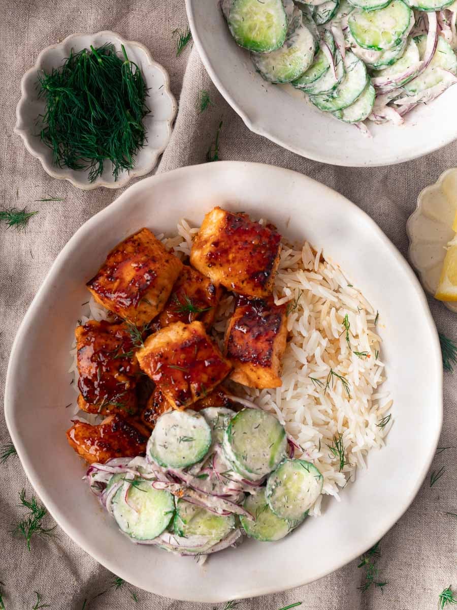 Honey harissa salmon with white rice and cucumber salad in a bowl, garnished with fresh dill on a neutral tablecloth.