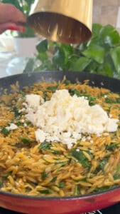 Orzo pasta with spinach and crumbled feta in a pan, topped with grated cheese.