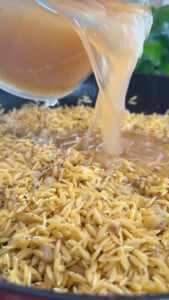 Pouring broth into a pan of seasoned orzo pasta.
