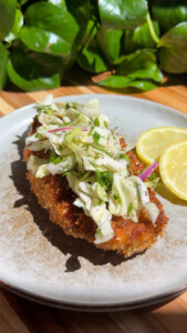 Crispy chicken cutlet topped with coleslaw and lemon slices on a plate, surrounded by green foliage.