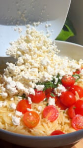 Feta cheese being added to orzo pasta with cherry tomatoes in a white bowl.