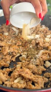 Hand adding spices to sautéed onions and ground meat in a pan.