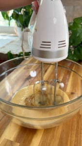 Hand mixer blending batter in a glass bowl on wooden countertop.