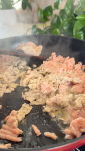 Sizzling ground meat cooking in a frying pan with steam rising, surrounded by green plants in the background.
