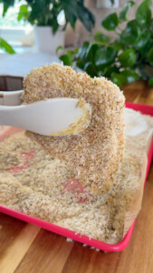 Tongs holding breaded chicken cutlet over a tray of breadcrumbs in a kitchen setting.