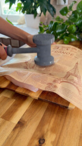 Person tenderizing chicken with a meat mallet on a wooden cutting board, covered with paper.