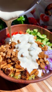 Chopped salad bowl with grilled chicken, cucumbers, tomatoes, and tahini dressing being poured on top.
