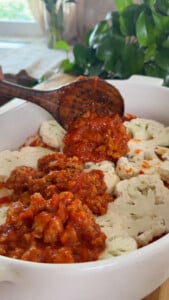 Wooden spoon serving tomato meat sauce over cauliflower slices in a white dish.