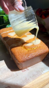 Pouring glaze over a loaf of golden-brown cake on a wooden table.