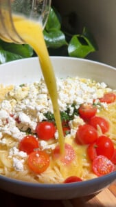 Pouring dressing over orzo salad with cherry tomatoes and feta cheese in a white bowl.