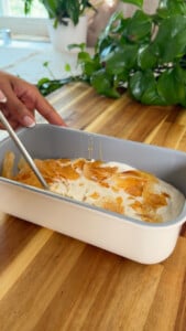 Hand preparing homemade ice cream with phyllo flakes in a white container on a wooden countertop.