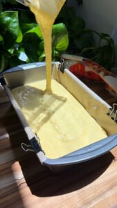 Pouring yellow batter into a parchment-lined loaf pan on a wooden table.