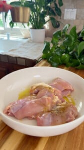 Raw chicken thighs with seasoning in a white bowl on a wooden countertop.