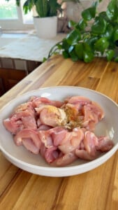 Raw chicken pieces with spices on a white plate in a kitchen setting.