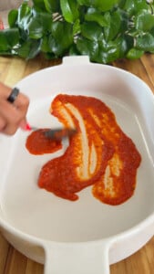 Spreading red tomato sauce on a white baking dish.