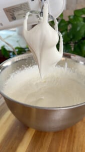Mixer with whipped cream peaks in a bowl on a wooden countertop.