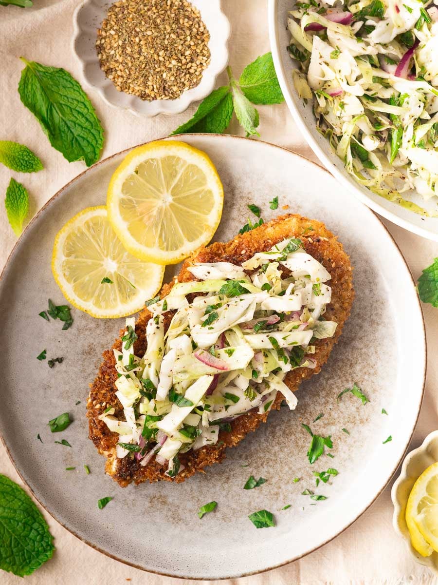 Za'atar chicken cutlet with Lebanese cabbage salad topped with fresh cabbage slaw and lemon slices on a plate, surrounded by herbs and spices.