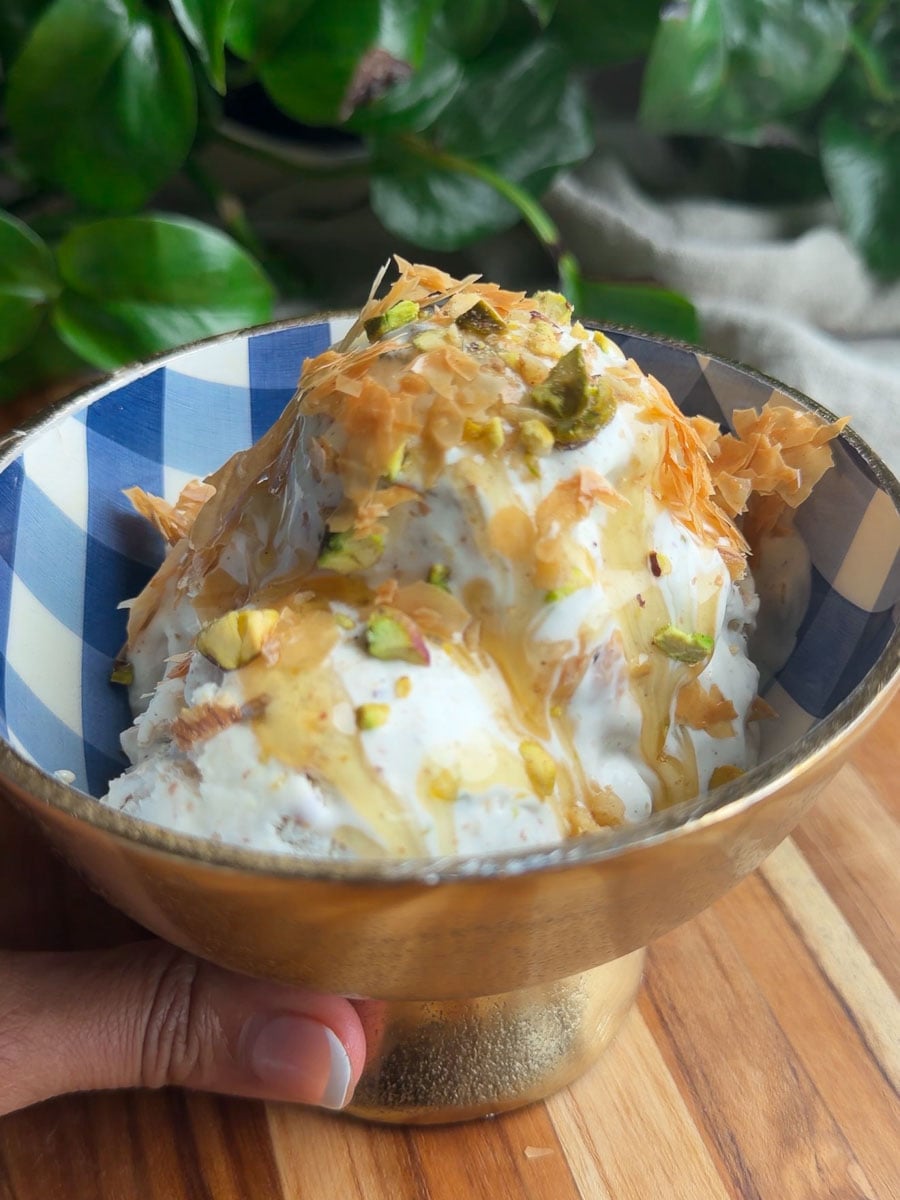 No-churn baklava ice cream topped with pistachios, phyllo pastry, and honey in a blue and white bowl on a wooden surface.