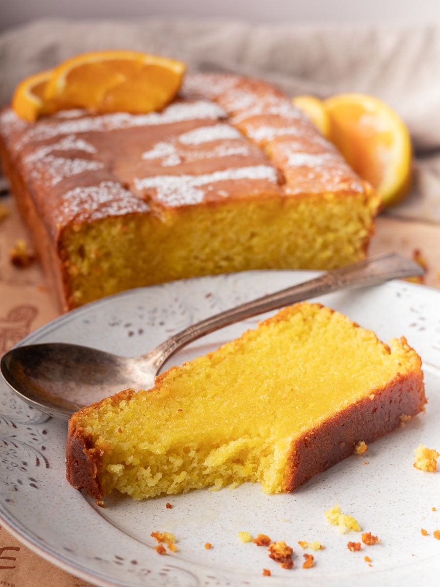 Slice of orange blender cake with powdered sugar on plate, silver spoon beside it, garnished with an orange wedge.