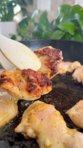 Chicken being cooked in a pan, with a piece held by tongs.