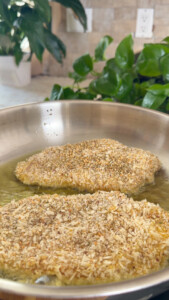 Breaded chicken frying in a pan with green plants in the background.