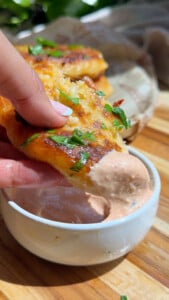 Hand dipping cheesy tuna cake with herbs into creamy sauce in a white bowl.