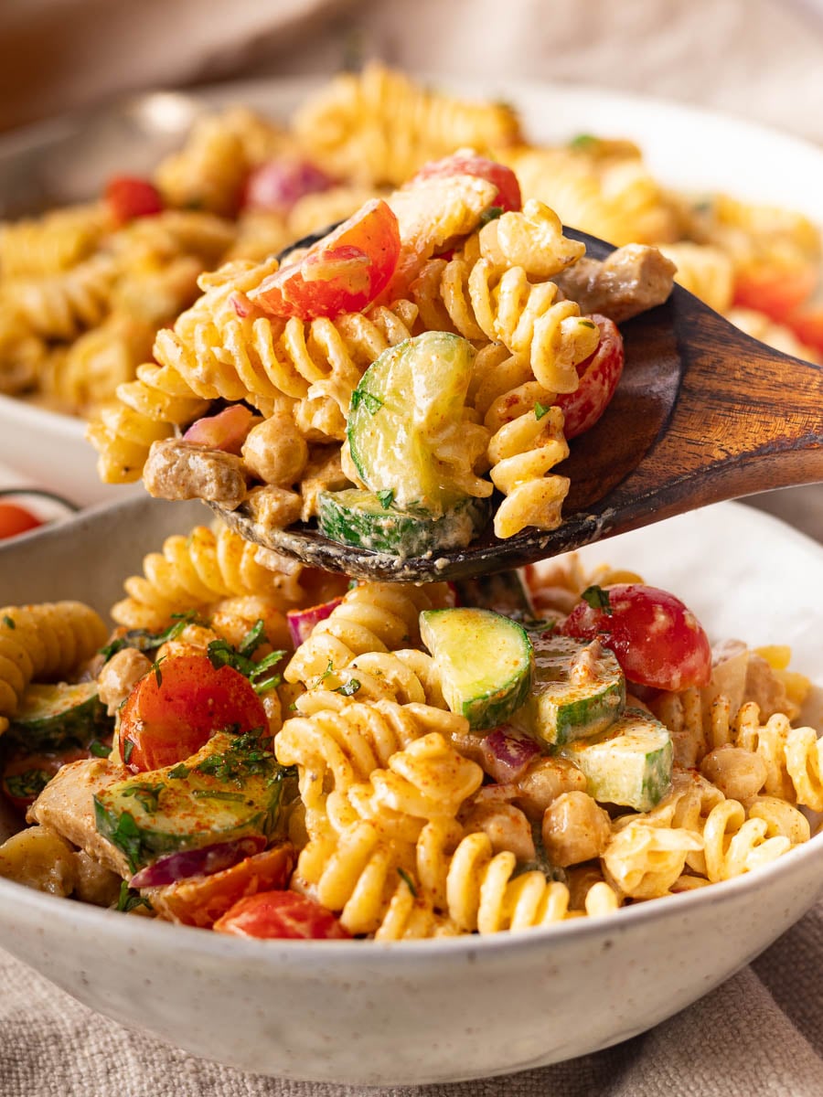 Mediterranean pasta salad with fusilli, cherry tomatoes, cucumber, and herbs on a wooden spoon in a white bowl.