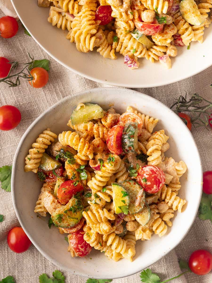 Two bowls of creamy Mediterranean pasta salad with cherry tomatoes, cucumbers, and herbs on a beige tablecloth.