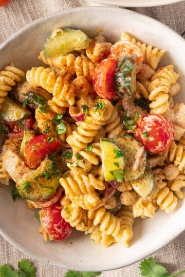 Mediterranean pasta salad with cherry tomatoes, zucchini, herbs, and creamy dressing on a textured cloth background.