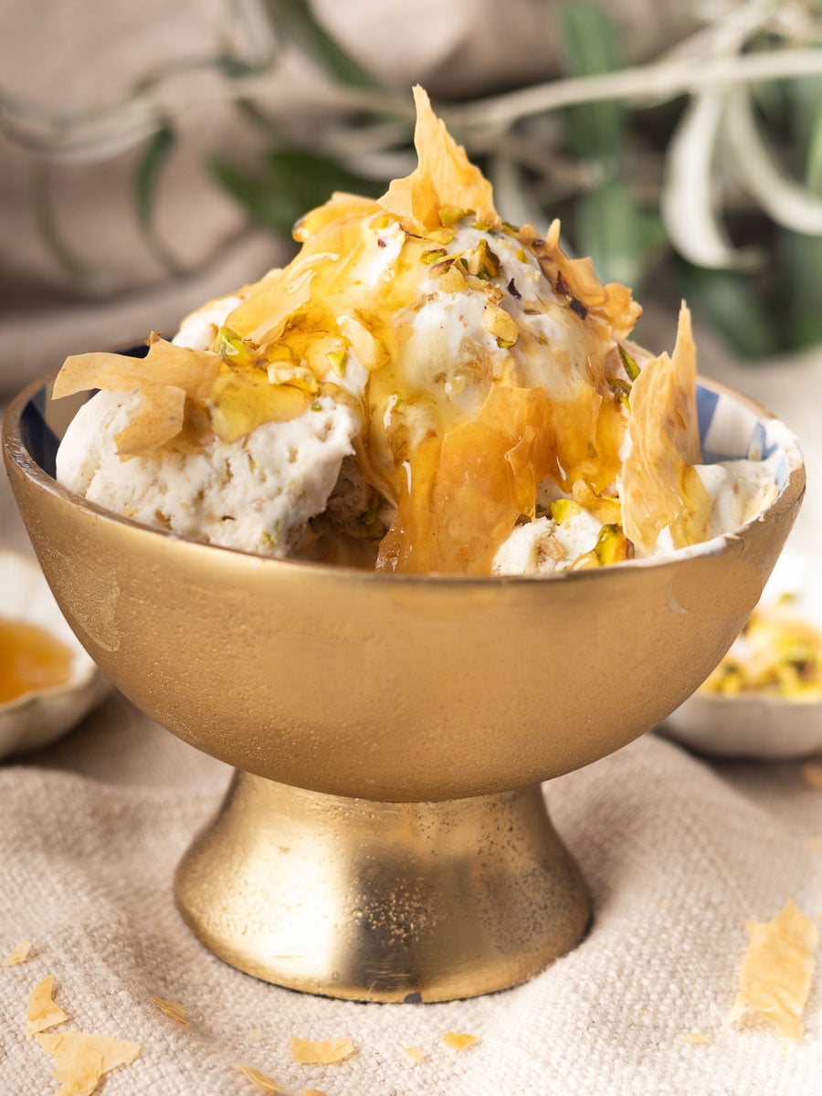 Bowl of no-churn baklava ice cream, topped with honey, pistachios, and pastry, on a textured beige cloth background.