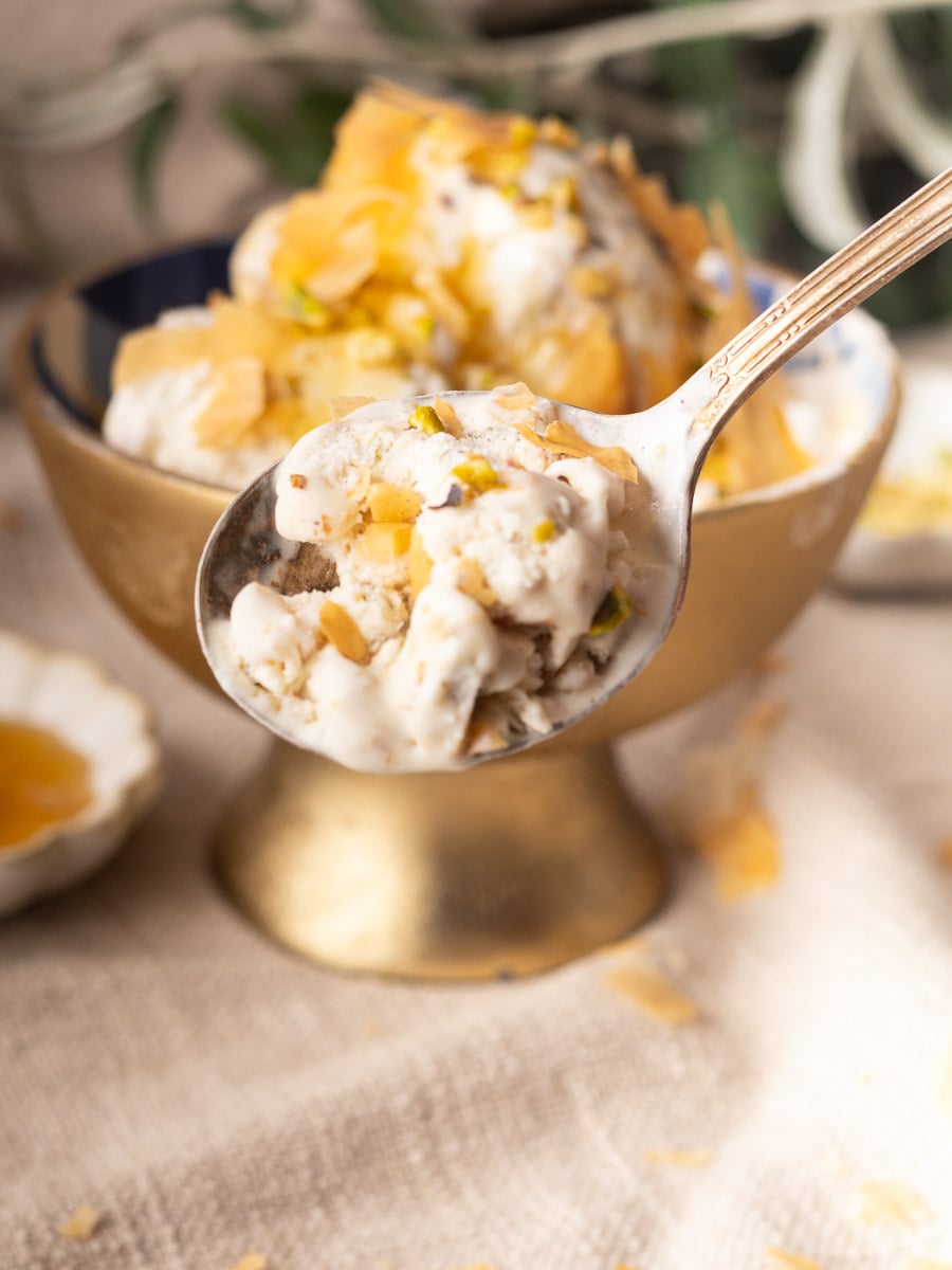 Spoon of no-churn baklava ice cream, with nut toppings in a gold bowl on a textured surface.